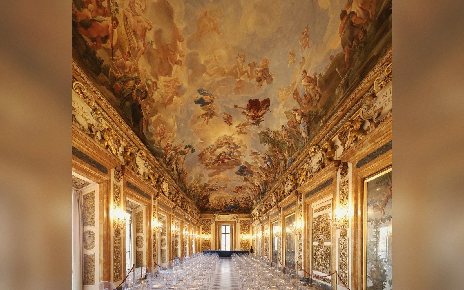 A gilded baroque gallery with an elaborate frescoed ceiling and a long banquet table set with glassware